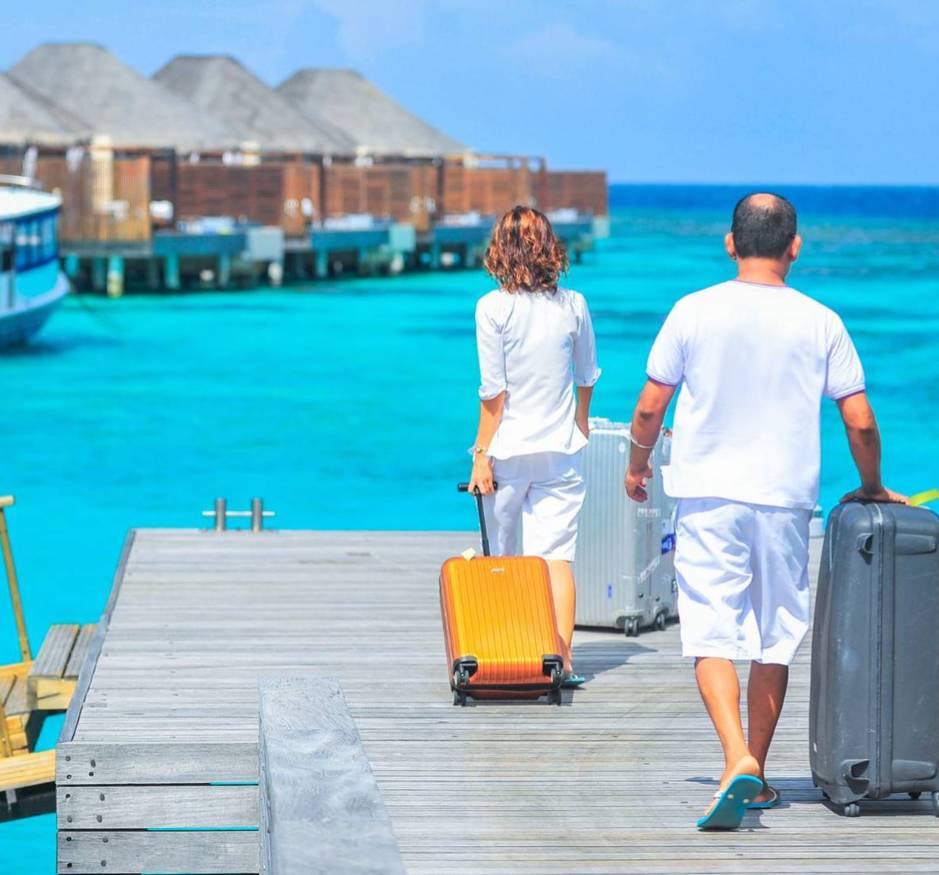 Two people with suitcases walking away from the viewer down a dock. A building is visible out on stilts over an azure sea.